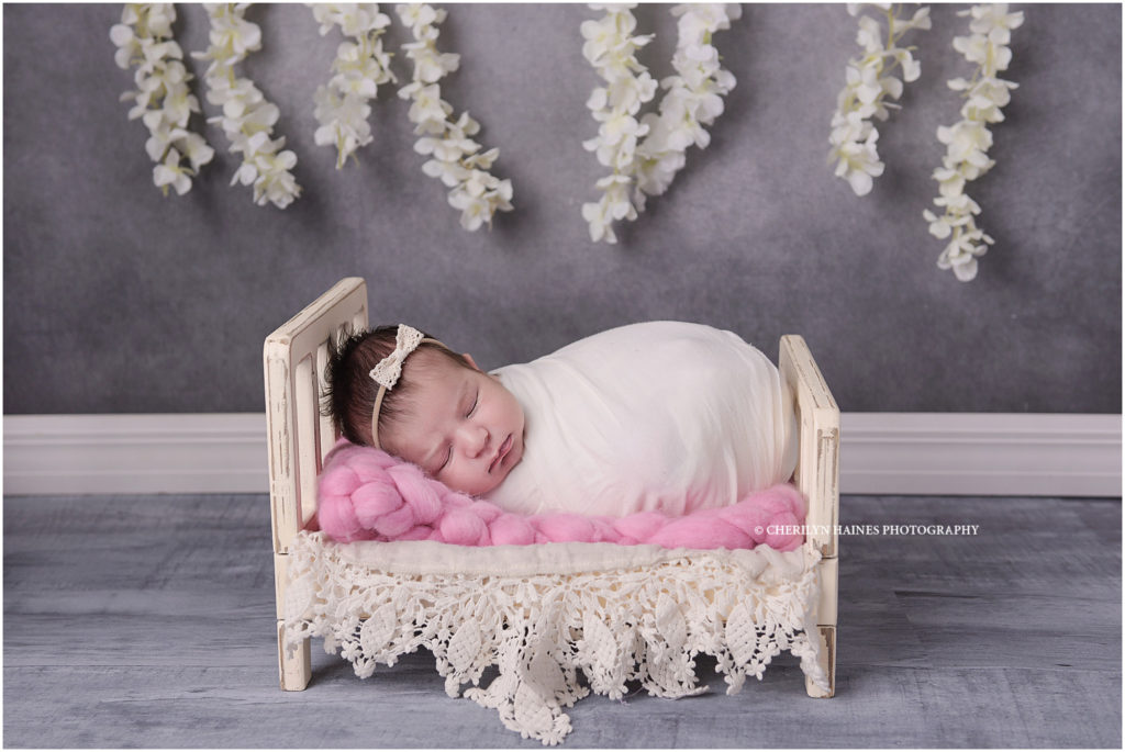 23 day old newborn baby girl photographed in tiny bed with florals hanging in the background
