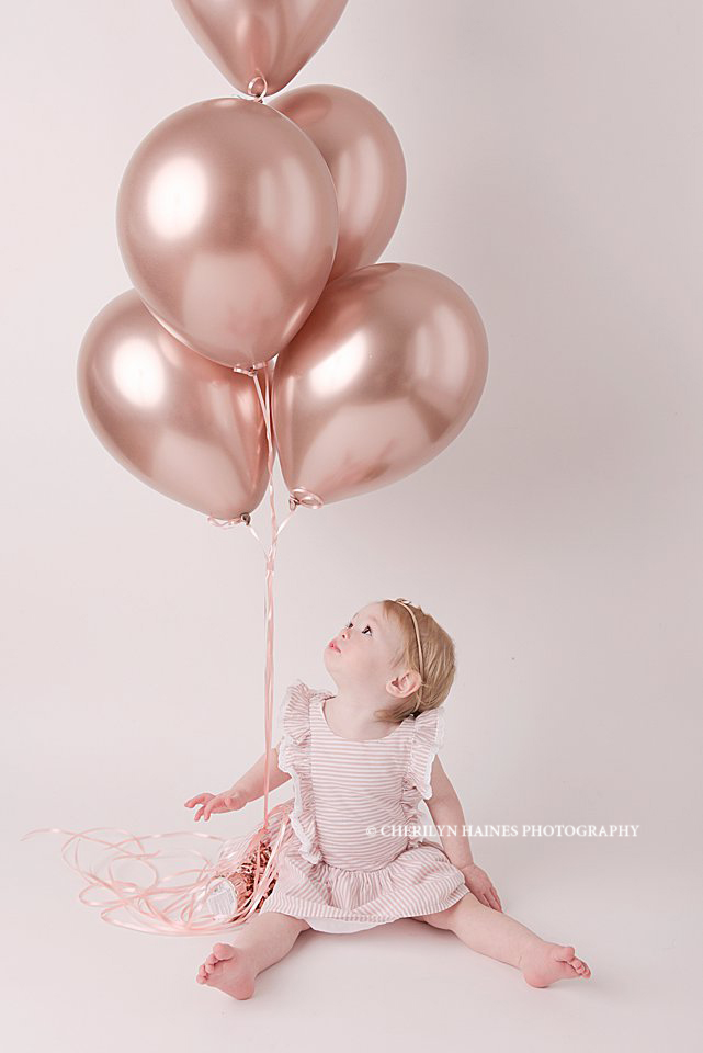 1 year old baby girl photographed looking up at rose gold balloons; baby session with Cherilyn Haines Photography