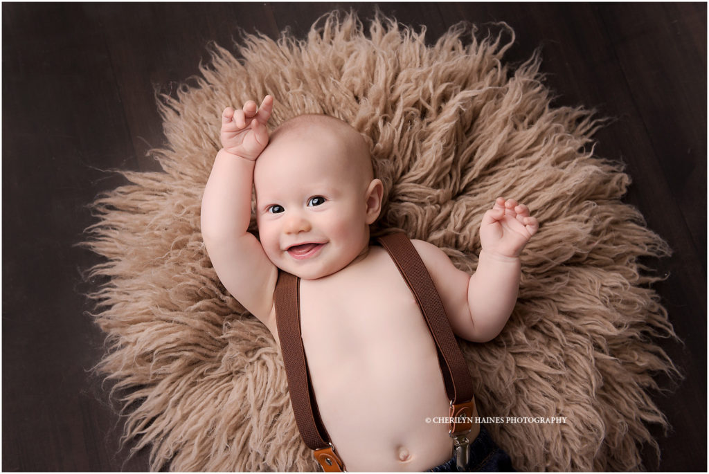 portrait of a 6 month old baby boy photographed by cherilyn haines photography in baton rouge, louisiana; baby boy wearing brown suspenders laying on tan flokati rug on rustic wood floordorop