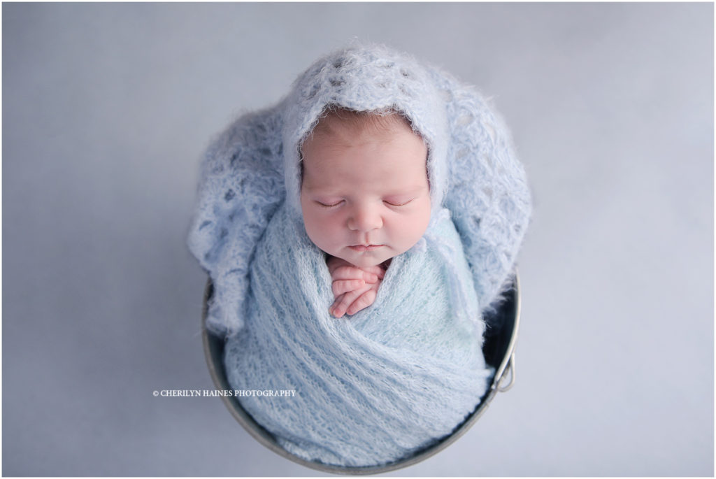 11 day old newborn baby girl photographed in a bucket on a blue backdrop while wearing a crocheted bonnet and swaddle; Cherilyn Haines Photography in Baton Rouge, Louisiana