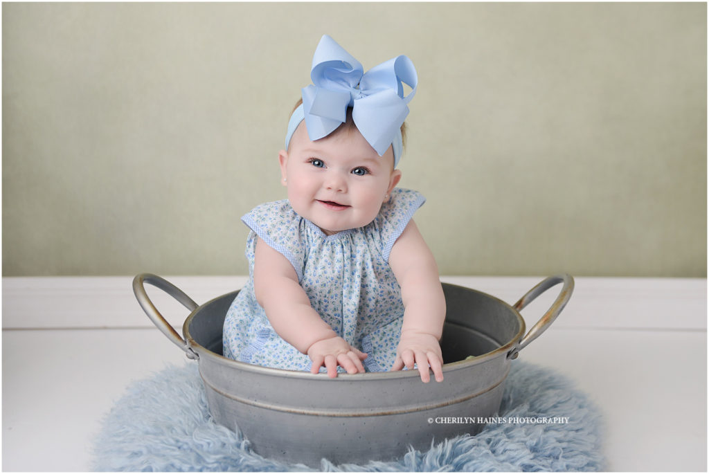 portrait of 6 month old baby girl wearing blue and green floral dress sitting in a metal bucket on green backdrop with light blue rug; photographed by cherilyn haines photography in baton rouge, louisiana