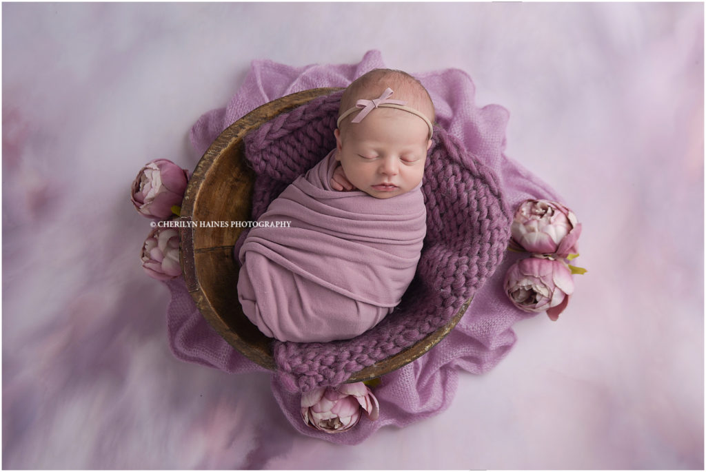 12 day old newborn baby girl photographed by cherilyn haines photography in baton rouge, louisiana; baby girl photographed swaddled in purple wrap on purple painted backdrop with lavender peonies