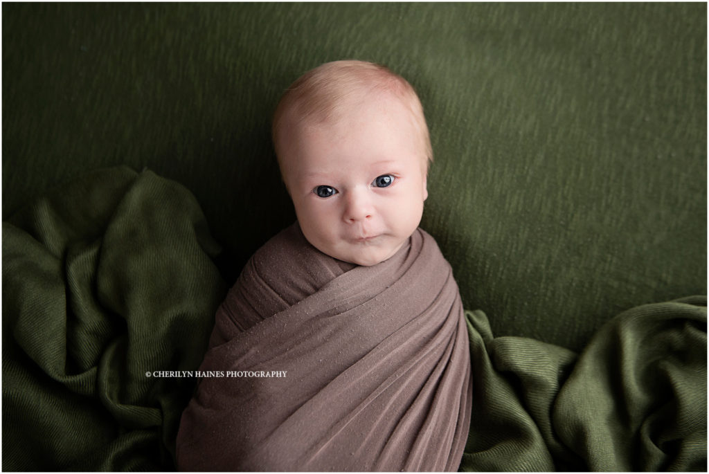 awake newborn baby boy photographed by cherilyn Haines photography in baton rouge, louisiana; baby boy swaddled in brown wrap laying on olive green blanket