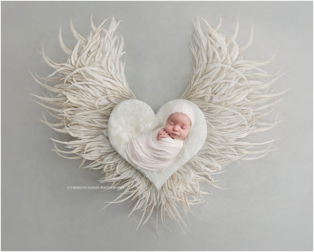 newborn baby girl photographed on white heart prop with angel wings