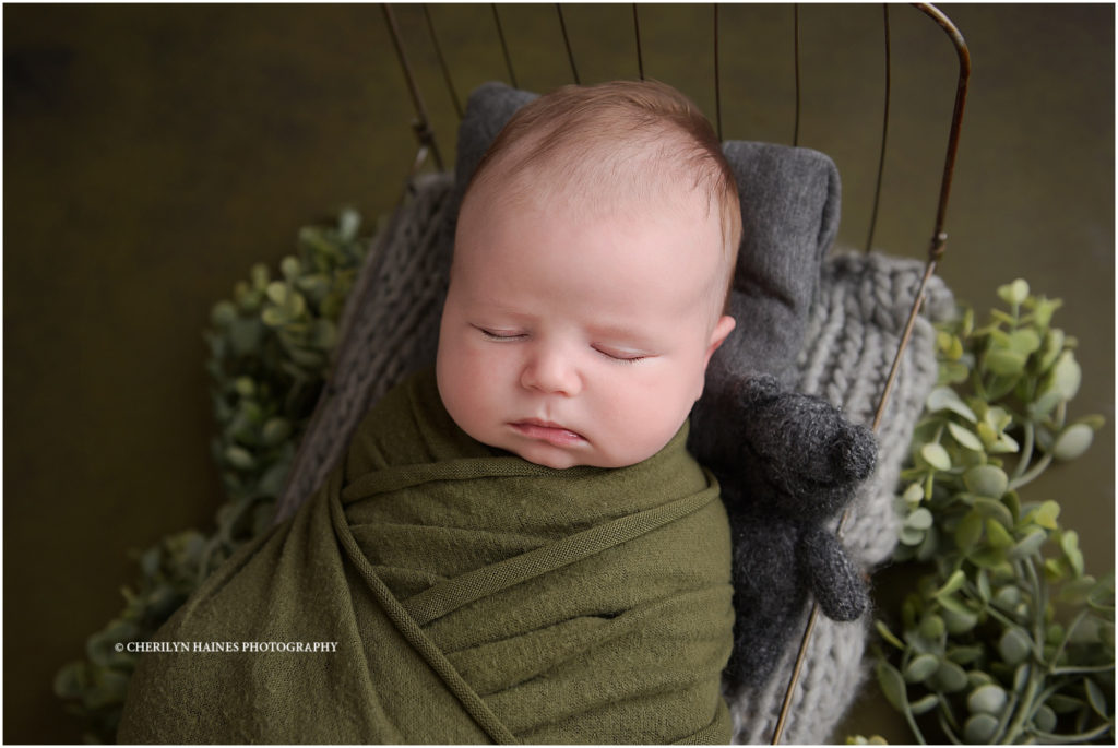 6 week old newborn baby boy photographed in baton rouge, louisiana; baby boy laying on tiny iron bed swaddled in olive green and holding a tiny teddy bear